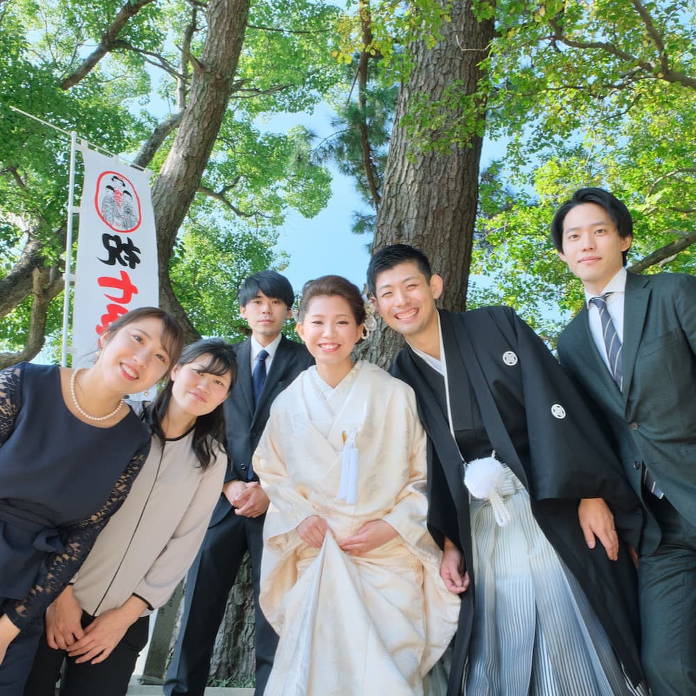 白無垢を着た花嫁様と紋付羽織袴を着た新郎様が神社で友人と笑っている