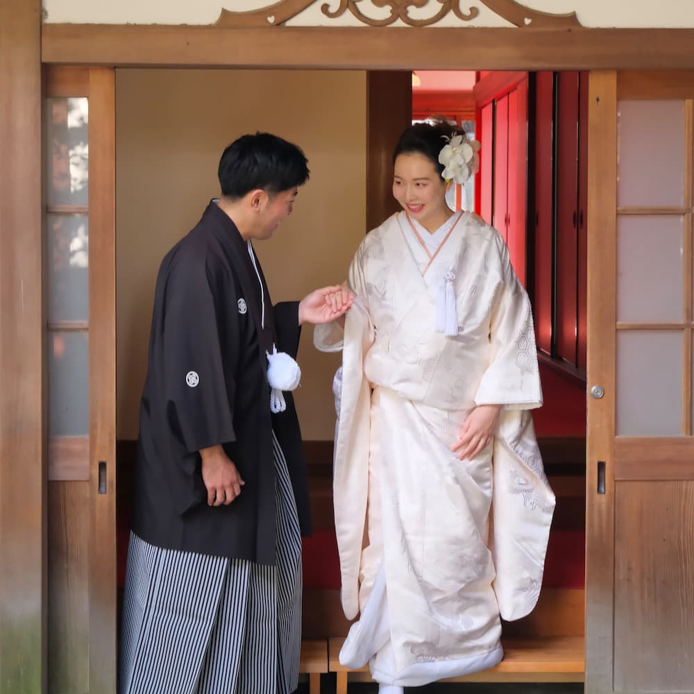 白無垢を着た花嫁様と紋付羽織袴を着た新郎様が神社で手を繋いで歩いている